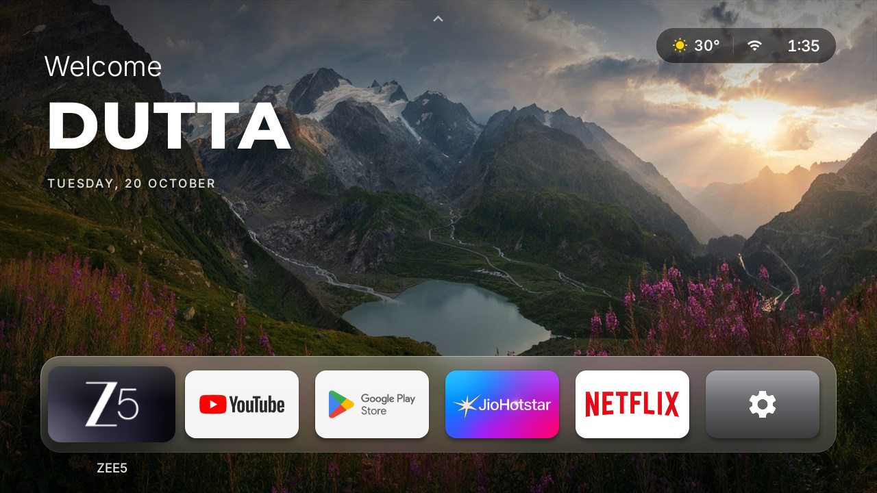 The cleanOS home screen on a TV: a mountain lake at sunset fills the screen, a large greeting with the date sits at top left, a weather, Wi-Fi and clock capsule at top right, and a frosted-glass dock holds six app tiles with the first one lifted and its name shown underneath.